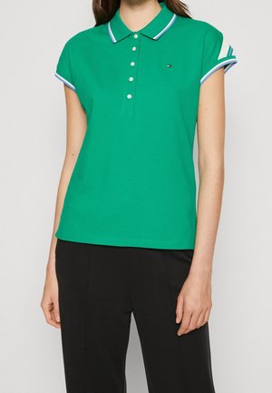 Femme portant un polo vert à manches courtes avec des boutons blancs et un col rayé, associé à un pantalon noir sur un fond uni.