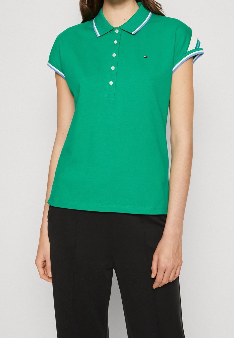 Femme portant un polo vert à manches courtes avec des boutons blancs et un col rayé, associé à un pantalon noir sur un fond uni.
