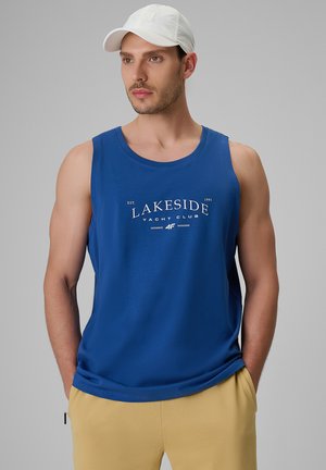 Mężczyzna w białej czapce, niebieskiej koszulce bez rękawów z tekstem "Lakeside Yacht Club" i żółtych spodniach, stoi z rękami w kieszeniach.