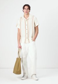 Uniformă compusă dintr-o cămașă cu mânecă scurtă, de culoare crem, cu modele geometrice maronii, asociată cu pantaloni albi cu croială largă și o geantă de umăr bej. Adidașii completează ținuta.