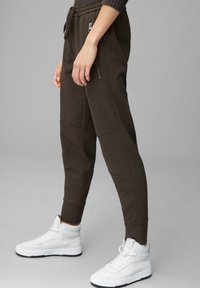 Braune Jogginghosen mit einem Kordelzugbund, seitlichen Reißverschlusstaschen und strukturiertem Stoff; kombiniert mit weißen High-Top-Sneakern.