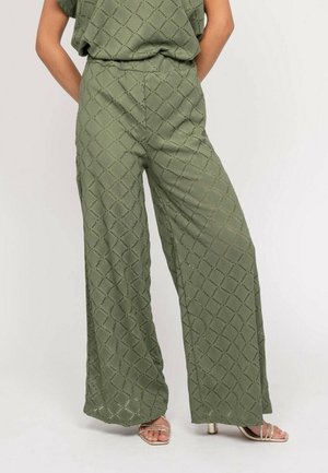 Femme portant un pantalon taille haute à jambes larges vert olive en tissu ajouré à motif diamant, avec des talons beige à brides.