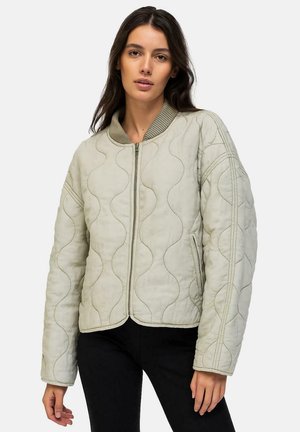 Vrouw met een lichtgrijze doorgestikte jas met een rits aan de voorkant, een geribde kraag en een zwarte broek tegen een effen achtergrond.