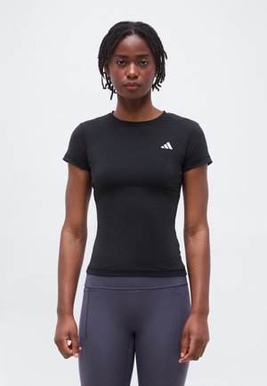 Γυναίκα με κοντά dreadlocks, φοράει μαύρο στενό μπλουζάκι Adidas και γκρι αθλητικό κολάν, στέκεται μπροστά σε ένα απλό φόντο.