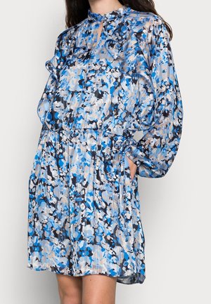 Femme portant une robe imprimée florale bleue, noire et beige avec de longues manches bouffantes et des détails à volants, main dans la poche.