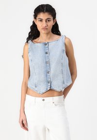 Chaleco de denim azul claro recortado con un escote recto, cinco botones en la parte delantera y un dobladillo en punta. Llevarlo sobre pantalones blancos de talle alto.