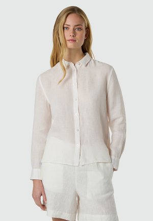 Giovane donna con capelli biondi lunghi che indossa una camicia bianca trasparente con bottoni e pantaloncini bianchi abbinati, su uno sfondo semplice.