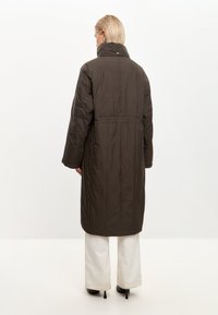 Persona con capelli biondi corti che indossa un lungo cappotto imbottito marrone scuro, pantaloni bianchi a gamba larga e scarpe nere con tacco alto, vista di spalle.
