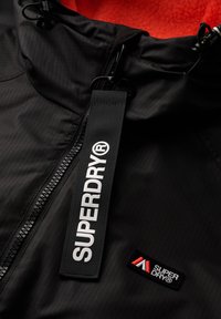 Chaqueta negra con sutil textura acanalada y forro de felpa roja. Presenta un logo blanco prominente y una marca con acentos rojos. Cierre de cremallera con tirador.