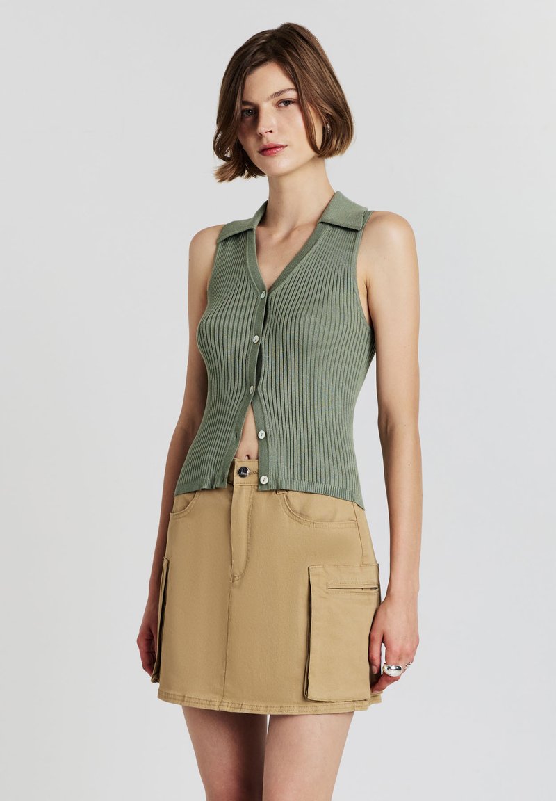 Giovane donna che indossa un top verde a coste senza maniche e una mini gonna cargo beige con tasche, in piedi su uno sfondo bianco.