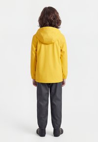 Bambino di spalle che indossa un impermeabile giallo con cappuccio, pantaloni impermeabili scuri e stivali da pioggia neri su sfondo bianco.