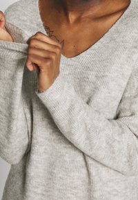 Jasnoszary sweter z dekoltem w kształcie litery V, o prążkowanej strukturze i długich rękawach; materiał wydaje się miękki i przytulny, z subtelnymi wariacjami kolorystycznymi.