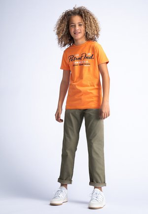 T-shirt in cotone arancione con testo grafico nero, abbinato a pantaloni verdi oliva con risvolto e scarpe bianche. Sfondo chiaro.