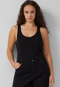 Zwarte tanktop met brede banden, ronde hals en aansluitende snit; gedragen met zwarte hoge taillebroek met zakken.