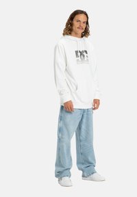 Biała bluza z logo na przodzie, w połączeniu z jasnoniebieskimi luźnymi dżinsami i białymi sneakersami. Materiał wydaje się miękki, przypominający bawełnę.