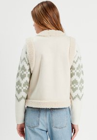 Fleece-Weste in Creme mit strukturiertem Kunstfellkragen und grünem geometrischen Muster an den Ärmeln, kombiniert mit Hellblauen Jeans.