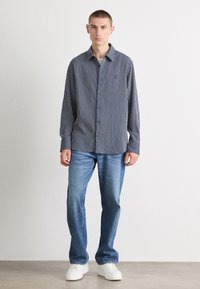 Ριγέ πουκάμισο με κουμπιά σε ναυτικό και λευκό, συνδυασμένο με ανοιχτό μπλε τζιν και λευκά αθλητικά παπούτσια. Το πουκάμισο έχει καπέλο με μυτερά κολάρα και μακριά μανίκια.