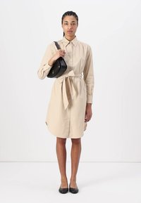 Abito camicia beige con vita a sbieco, maniche lunghe e colletto. Abbinato a una piccola borsa a spalla nera e scarpe nere piatte.