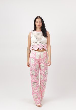 Mujer de pie con top corto de crochet blanco y pantalones de crochet con estampado floral rosa y blanco sobre un fondo liso.