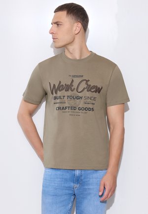 Mand iført olivengrøn t-shirt med teksten "Work Crew" og lyseblå jeans, kigger til venstre mod en hvid væg.