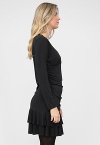 Schwarzes Langarmkleid mit gerafftem Oberteil, figurbetonter Silhouette und mehrlagigem Volant-Saum. Hergestellt aus weichem Stoff, geeignet für verschiedene Anlässe.