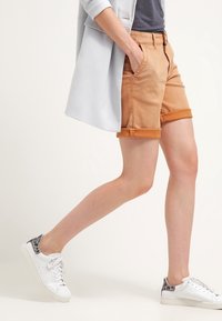 Shorts roulés de couleur marron clair avec des passants de ceinture et des poches latérales, associés à des baskets blanches avec un accent de motif serpent gris.