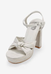 Beige suede high-heeled sandal with a knotted front strap, adjustable ankle strap, and a chunky platform sole.