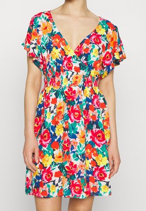 Kvinde iført en farverig kort blomsterkjole med V-hals og fine ærmer mod en ensartet lys baggrund.