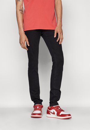 Vaqueros negros ajustados con una textura suave, combinados con zapatillas Nike rojas y blancas que presentan el distintivo logo de swoosh y un diseño clásico.