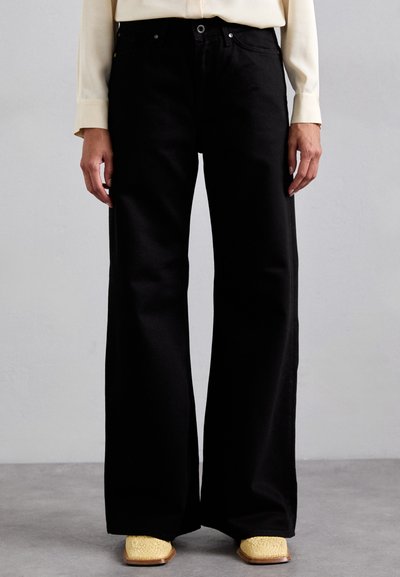 Pantalones de denim de gran ancho en color negro, con una cintura alta, detalles de costura sutiles y una textura suave. Complementados con zapatos tejidos amarillos.