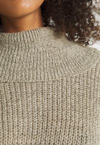 Vaaleanharmaa neulottu pusero, jossa on korkea ribbineuloskaulus. Puserossa on tekstuuri viimeistely ja väljähkö istuvuus, korostaen sen mukavaa muotoilua.