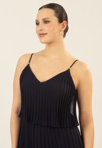Marineblaues Faltenkleid mit schmalen Trägern und goldenen Akzentdetails. Es hat einen V-Ausschnitt und ein mehrlagiges Design, das den strukturierten Stoff hervorhebt.