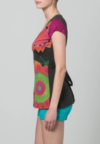 Barevný top s krátkými rukávy, který kombinuje živé vzory a jednobarevnou černou látku, s mašlí na pase. Nositelný s tyrkysovými krátkými kalhotami.