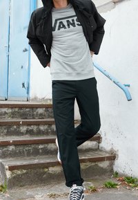 Schwarze Jacke, grauer Sweatshirt mit "VANS"-Logo, schwarze Hose und schwarze Sneaker mit weißen Akzenten. Außensetting mit blauer Tür und Treppe.