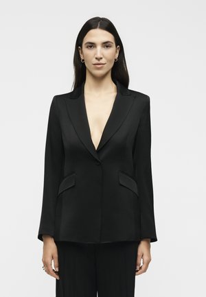 Γυναίκα με μακριά σκούρα μαλλιά, φοράει χρυσούς κρίκους στα αυτιά και ένα μαύρο, ραμμένο σακάκι με βαθύ ντεκολτέ, που στέκεται μπροστά σε απλό φόντο.