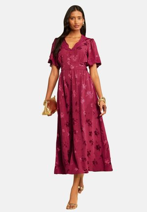 Vrouw draagt een lange, korte mouwen, diep roze bloemige jurk, houdt een gouden clutch vast en draagt gouden sandalen en armbanden.