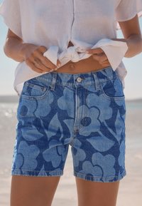 Siniset denimshortsit, joissa on kukkakuosi eri sinisen sävyissä, viiden taskun malli ja napitus vyötäröllä.
