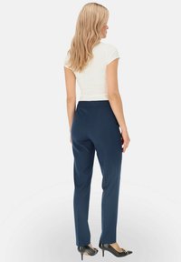 Granatowe spodnie garniturowe o dopasowanym kroju, gładkiej tkaninie i z pasem w talii. Noszone z czarnymi szpilkami. Widok z tyłu.