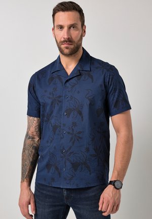 Mand med skæg og tatoveret arm iført en mørkeblå kortærmet blomsterknap-skjorte og mørke jeans, stående mod en ensfarvet baggrund.