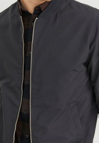 Ciemnoszara kurtka bomber z zamkiem błyskawicznym z przodu, ściągaczem przy kołnierzu i kieszeniami po bokach. Nosi się ją na brązowo-czarną koszulę w kratę.