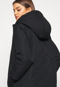 Veste noire matelassée avec capuche, arborant un motif en losanges et des poches latérales. Le tissu est lisse avec une finition mate.