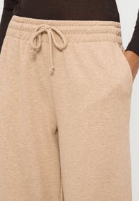 Gros plan sur un pantalon de survêtement beige avec taille à cordon et poche latérale, porté avec un haut à manches longues foncé.