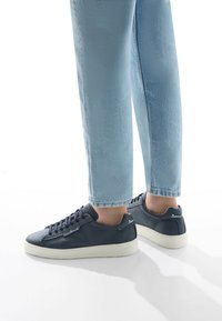 Sneakers in pelle navy con superficie testurizzata, suola in gomma bianca e chiusura con lacci, abbinati a jeans azzurri chiari.