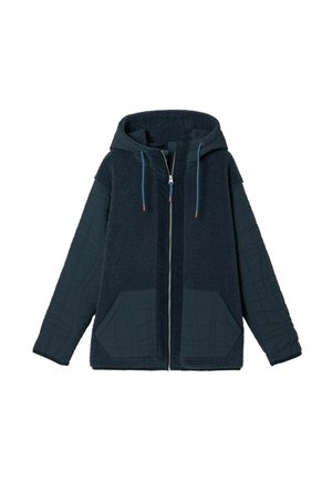 Kvinde iført navyblå quiltet og fleecet jakke over en stribet skjorte og blå jeans, stående med den ene hånd i lommen mod en ensfarvet baggrund.