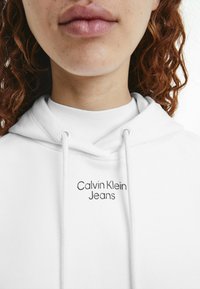 Sudadera blanca de algodón con capucha, que cuenta con cordones y el logotipo de "Calvin Klein Jeans" en texto negro bordado en el pecho.