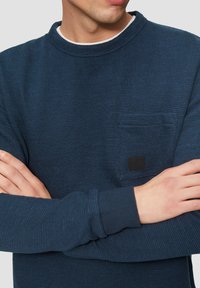 Marineblaues Strick-Sweatshirt mit dezenten blauen Streifen, Rundhalsausschnitt, langen Ärmeln und einer kleinen Brusttasche mit einem schwarzen Logo-Patch.