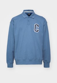 Sweat-shirt polo en coton bleu avec un col et des poignets côtelés, arborant un grand emblème blanc en forme de lettre "C" sur la poitrine gauche. Coupe classique.