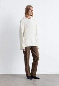 Cremfarbener, übergroßer Pullover mit geripptem Saum und Bündchen, kombiniert mit braunen, taillierten Hosen und schwarzen Slippers. Glatte Textur.