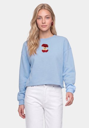 Junge Frau mit langen blonden Haaren, die ein hellblaues Sweatshirt mit einem roten Design eines angebissenen Apfelkerns und weiße Hosen vor einem einfarbigen Hintergrund trägt.
