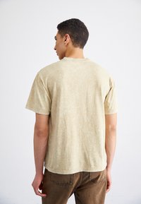 Camiseta de manga corta beige hecha de una tela texturizada, con un corte relajado y un cuello redondo, combinada con pantalones marrones.